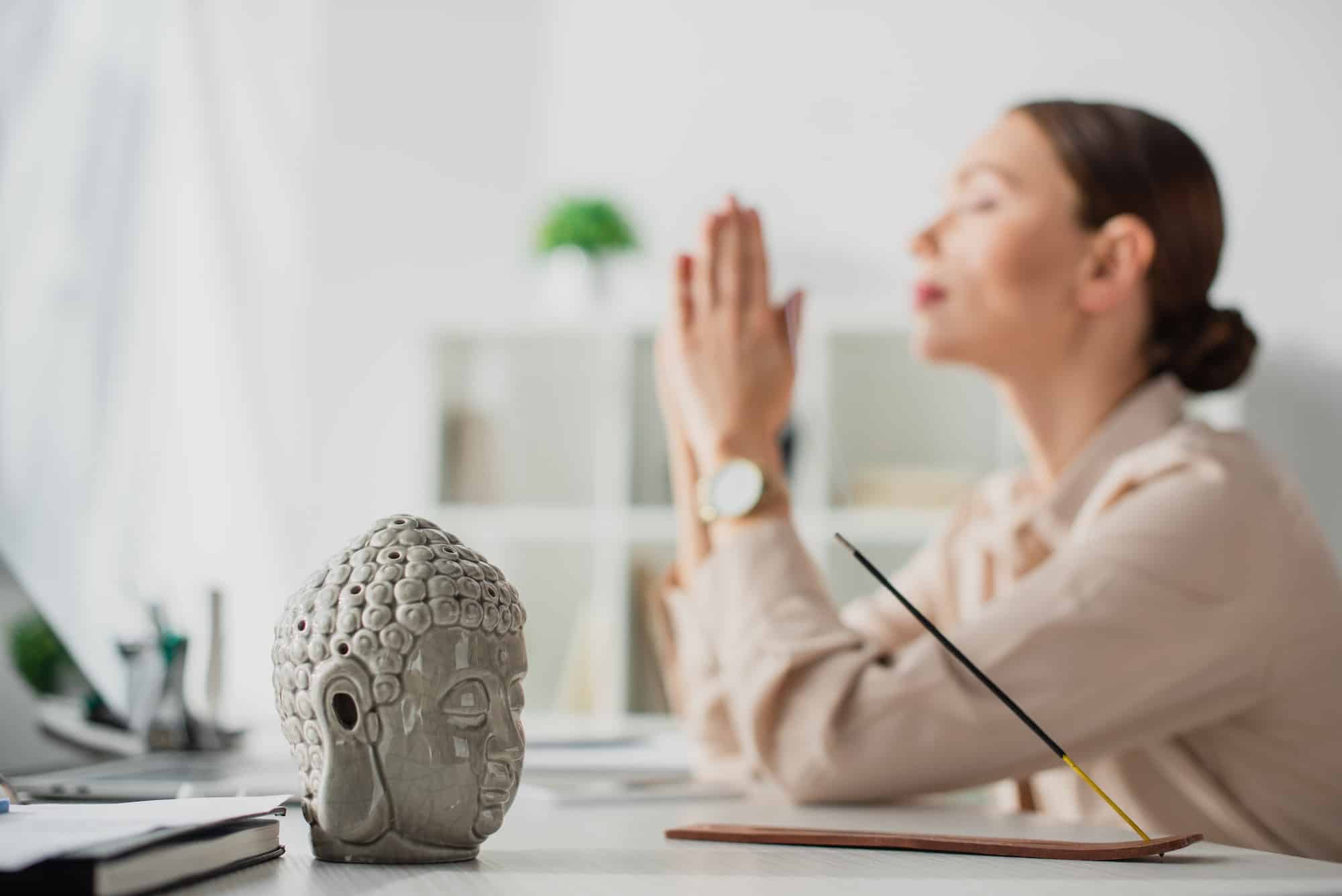 messa a fuoco selettiva di una donna d'affari che medita con il gesto di namaste sul posto di lavoro con la testa di Buddha e la testa di Buddha.